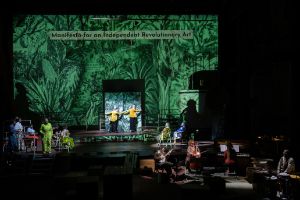 Schauspieler*innen auf der Bühne, davor Musiker*innen mit Instrumenten. Projektionen grüner Pflanzen im Hintergrund.